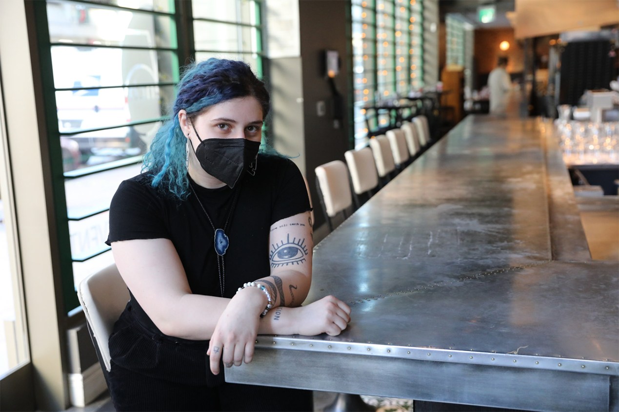 Nikki Perri sits at the end of long metal restaurant table. Behind her are empty chairs and turned over glasses.