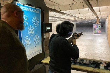 Russell Lewis stands behind Sharis Lewis at a dimly lit gun range as she braces a long gun against her shoulder, aiming at her target. The target is a sheet of paper printed with multicolored skulls labeled, "ransomeware dudes."