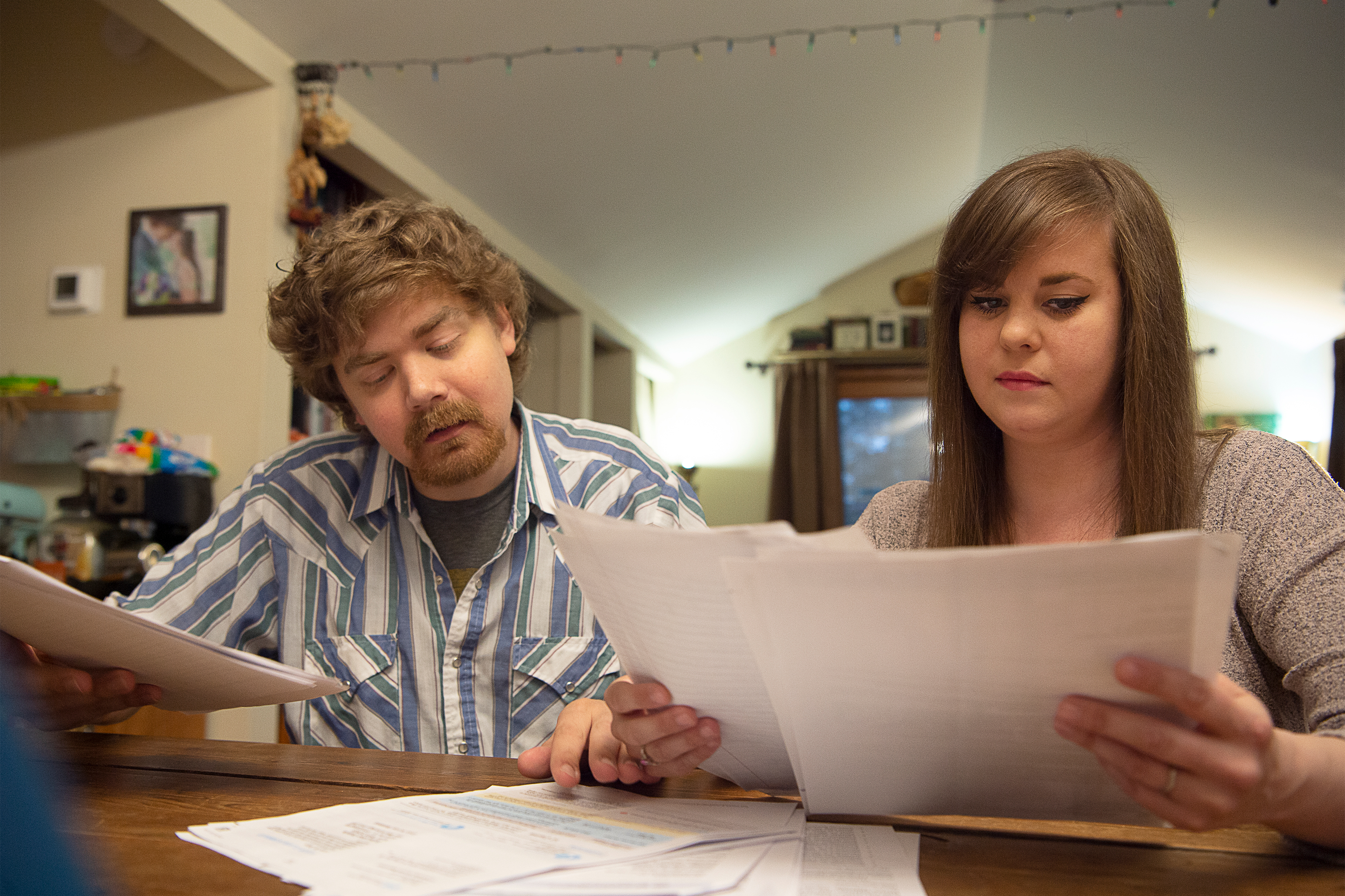 Sean Deines is seen sitting on the left side of a table with his wife, Rebekah, sitting to the right. The two are holding and looking over stacks of medical bills.