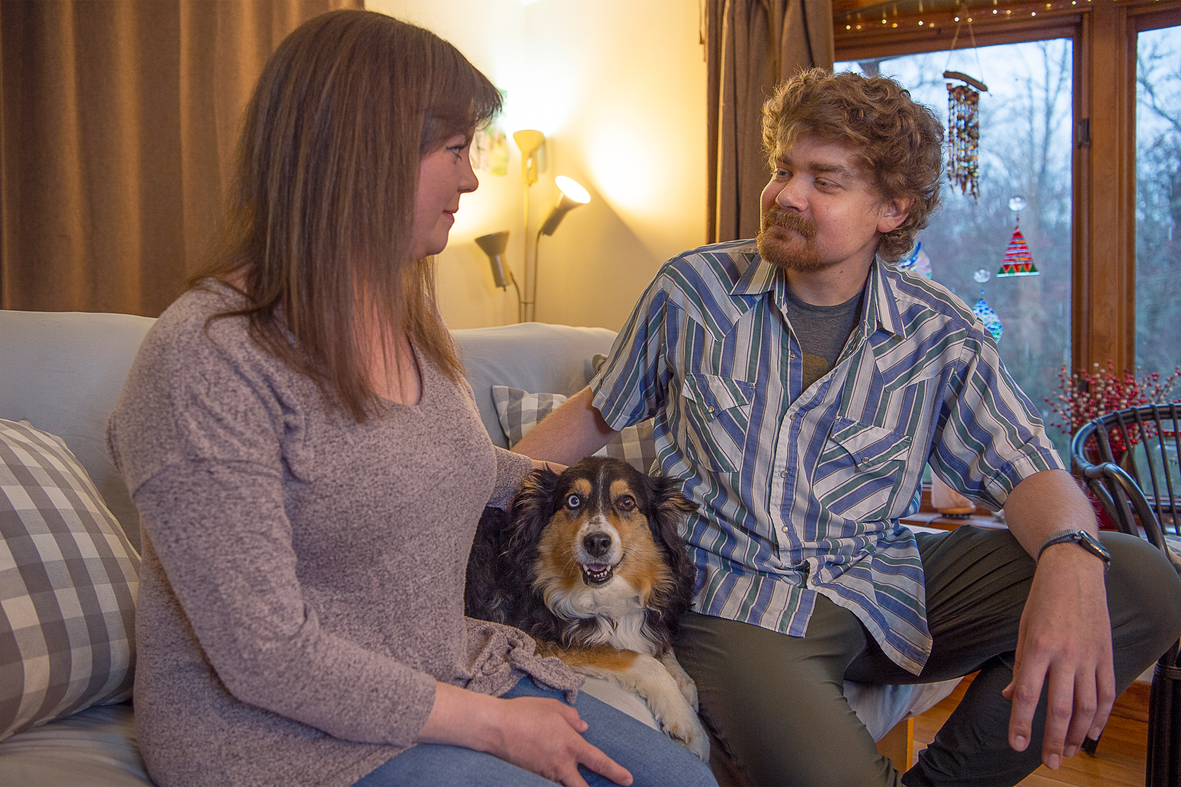 Rebekah Deines sits on the left side of a couch, smiling at her husband, Sean. Sean smiles back, sitting on the right, as their dog, Maddi, sits between them.