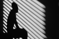 A photo of shadows on a wall shows the silhouette of a person sitting superimposed with the shadows of bars on a window behind them.