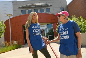 Two women are seen standing in front of a Planned Parenthood clinic wearing blue vests. The vests bear text that reads "Escort / Escorta."