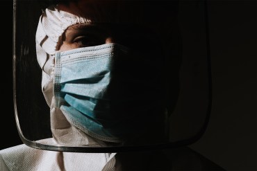 Harsh, angular shadows hide the right side of an ICU doctor's face. The doctor wears protective scrubs, a face mask and a plastic face shield.