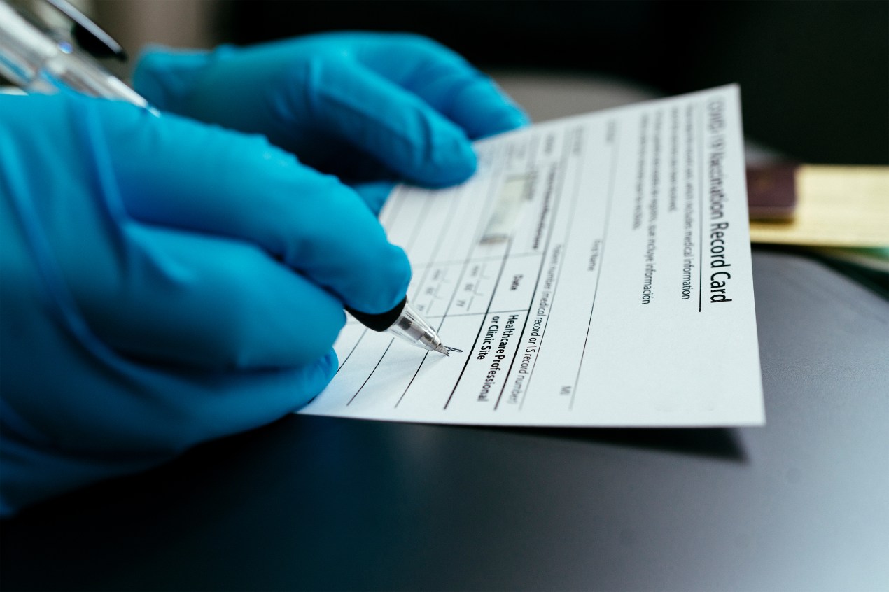 A pair of surgically gloved hands signs a covid-19 vaccination card.