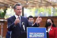 Gov. Gavin Newsom speaks from behind a podium, gesturing with his arms outside of a restaurant. A blue sign sits in front of the microphone at the podium that says, "The California Blueprint."