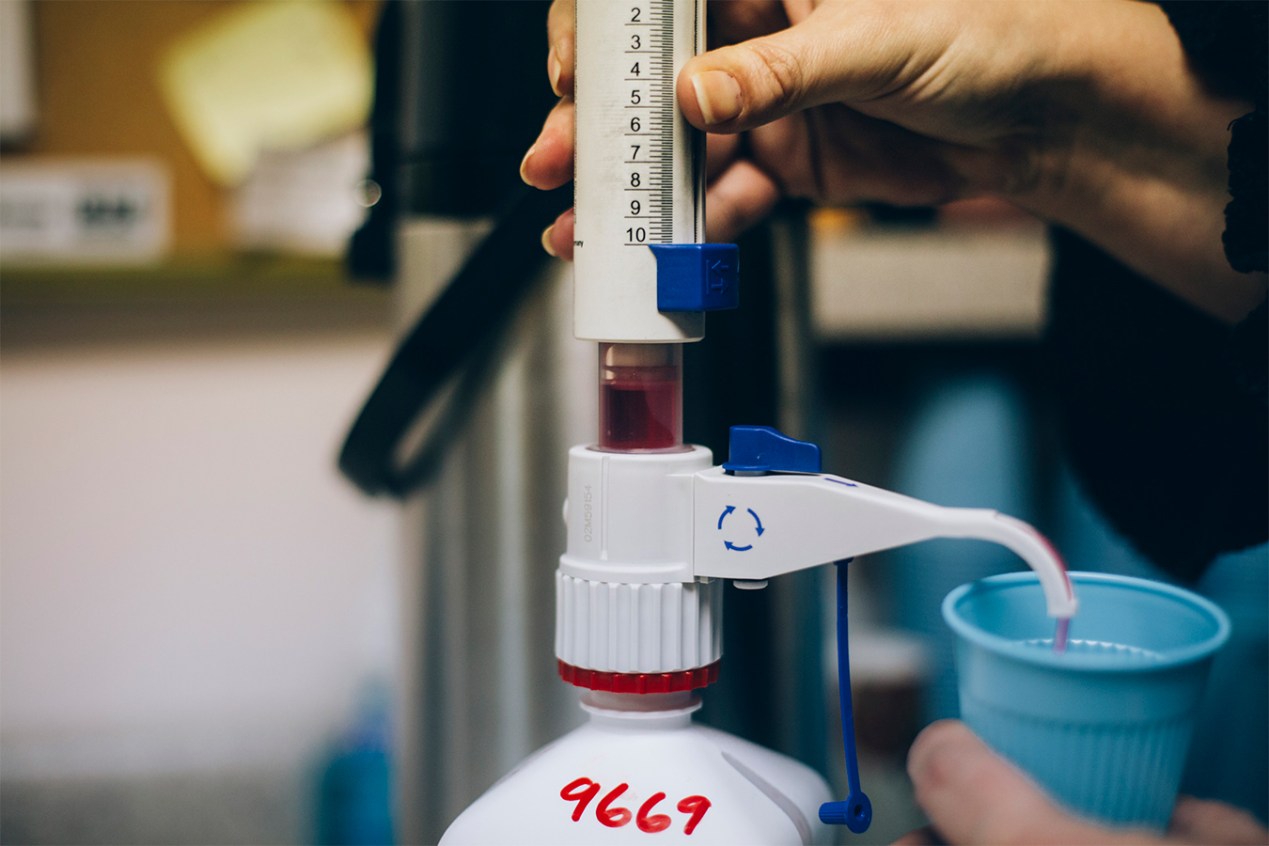 A dispenser pours a dose of methadone — a red liquid — into a plastic cup.