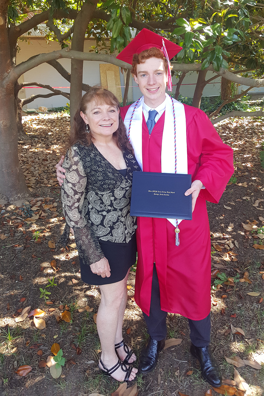 Tyler Gilreath stands next to his mother, Tamra, wearing a red cap and gown for his high school graduation, holding his diploma. His mother stands next to him on the left wearing a black patterned dress. They are standing outdoors and the weather is sunny.