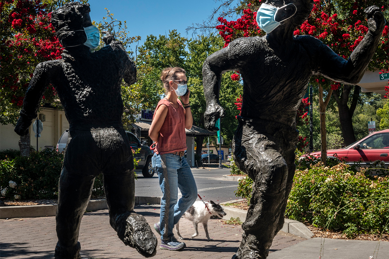 A woman is seen walking her dog on the sidewalk on a sunny day. She is framed on her left and right by two black statues of running children. Surgical masks are affixed across their faces.