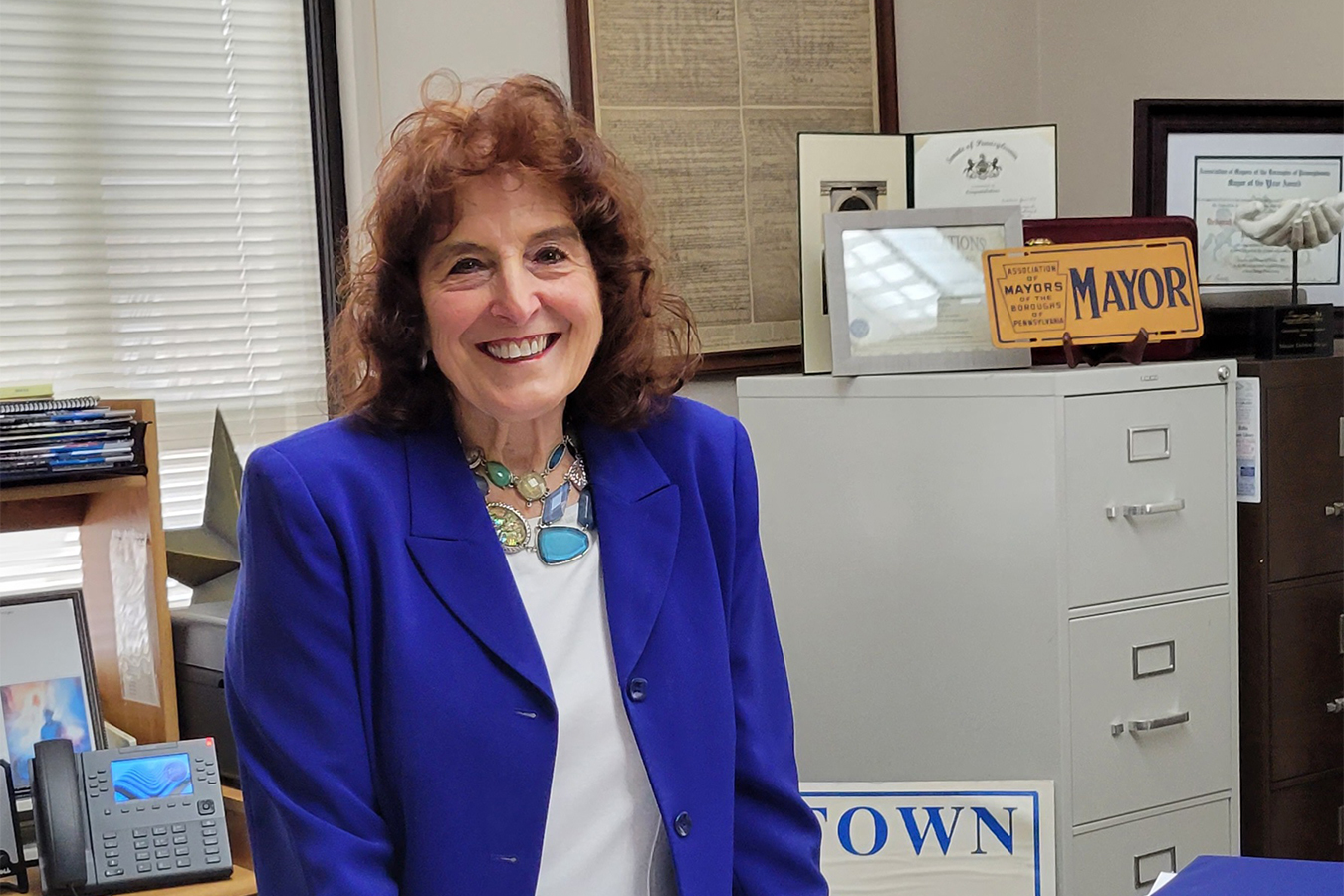 Mayor Deborah Bargo is seen standing in her office, smiling. A sign on top of filing cabinet reads, "Mayor," printed in bold text.
