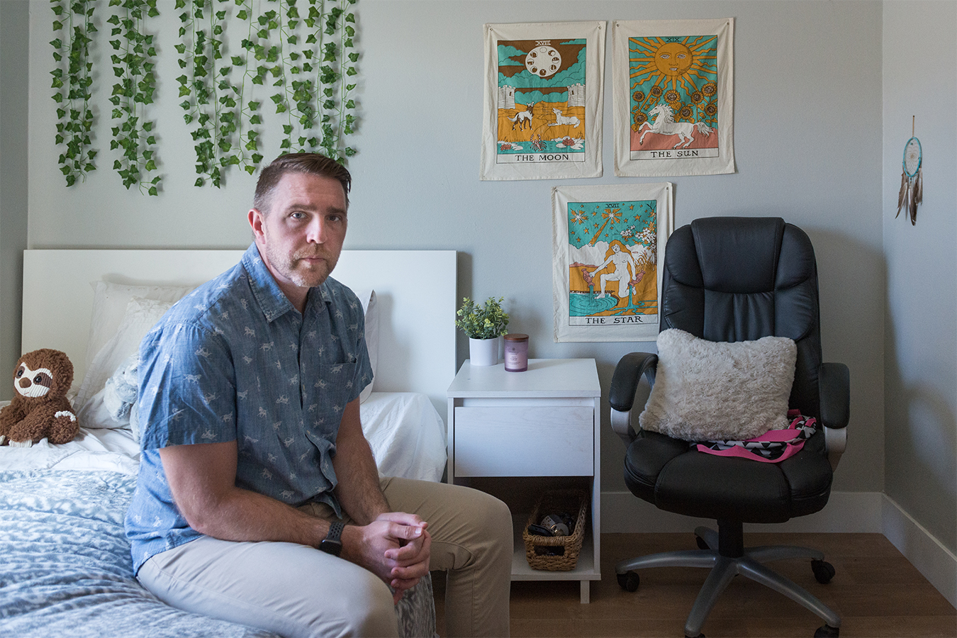 Lee Stonum is seen on the left, sitting in his daughter's bed. A plush sloth sits to his right. Canvas prints and ivy hang on the walls behind him. The room is brightly lit and the walls and furniture are white.