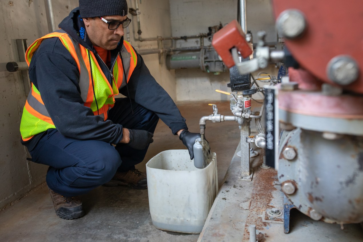 Patrick Green is seen on the left squatting and holding a bottle to a tap that siphons wastewaster. Excess sludge flows into a bucket underneath the tap.