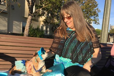 Diana McAllister is seen sitting on a park bench next to her dog, Honey. McAllister is wearing a turquoise shirt, Honey is wearing sunglasses, a visor and a skirt in the same color.