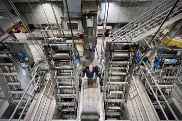 Pieter Van Ry is seen standing between two machines, smiling at the camera. He is seen from above, and a wide angle lens shows the facility around him.