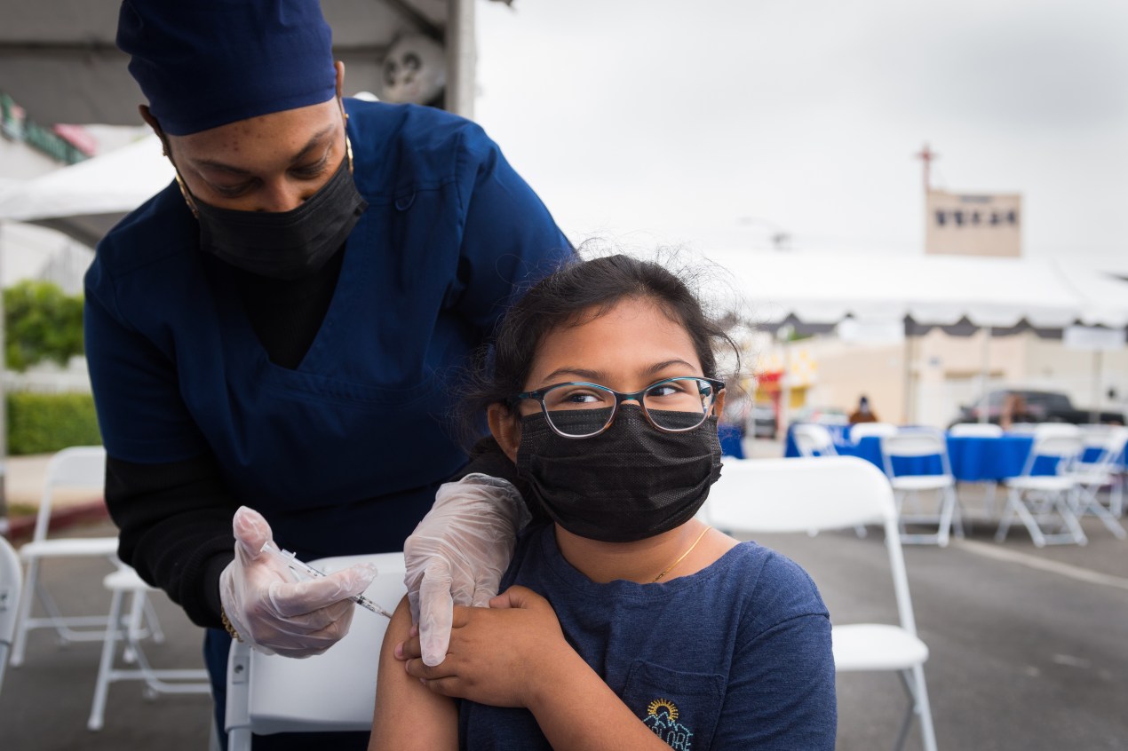A photo of a young girl, wearing a mask and glasses, getting vaccinated outdoors.