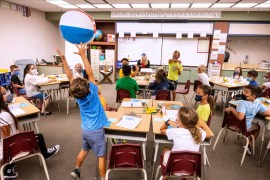 Children wearing masks are in a classroom taking part in a group activity. One child jumps up to catch a beach ball.