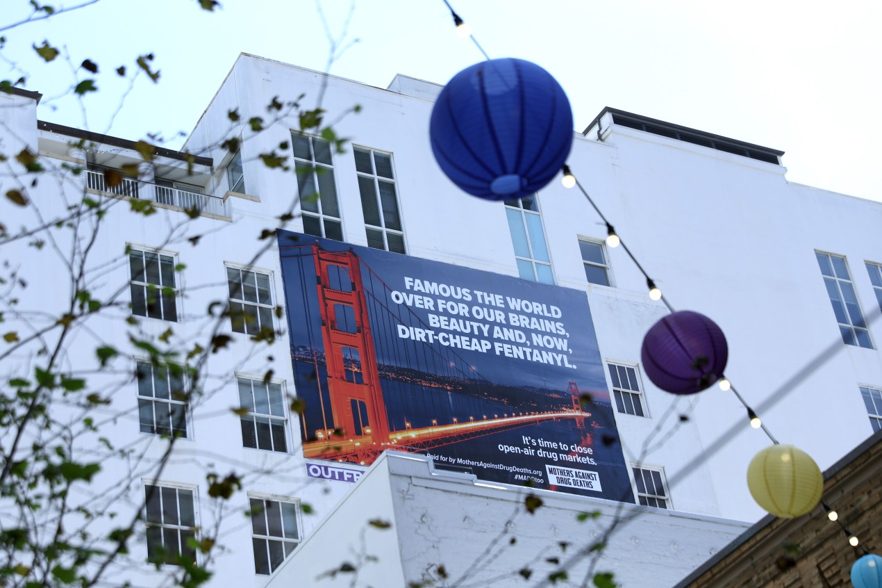 A billboard on the side of a building shows the Golden Gate Bridge with text next to it reading, "FAMOUS THE WORLD OVER FOR OUR BRAINS, BEAUTY AND, NOW, DIRT-CHEAP FENTANYL." Tree branches and a string of lanterns and lights are seen blurred in the foreground.