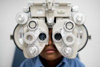 A closeup photo shows a child viewed from the front getting an eye exam with a phoropter. The large medical instrument obscures most of the child's face from view.