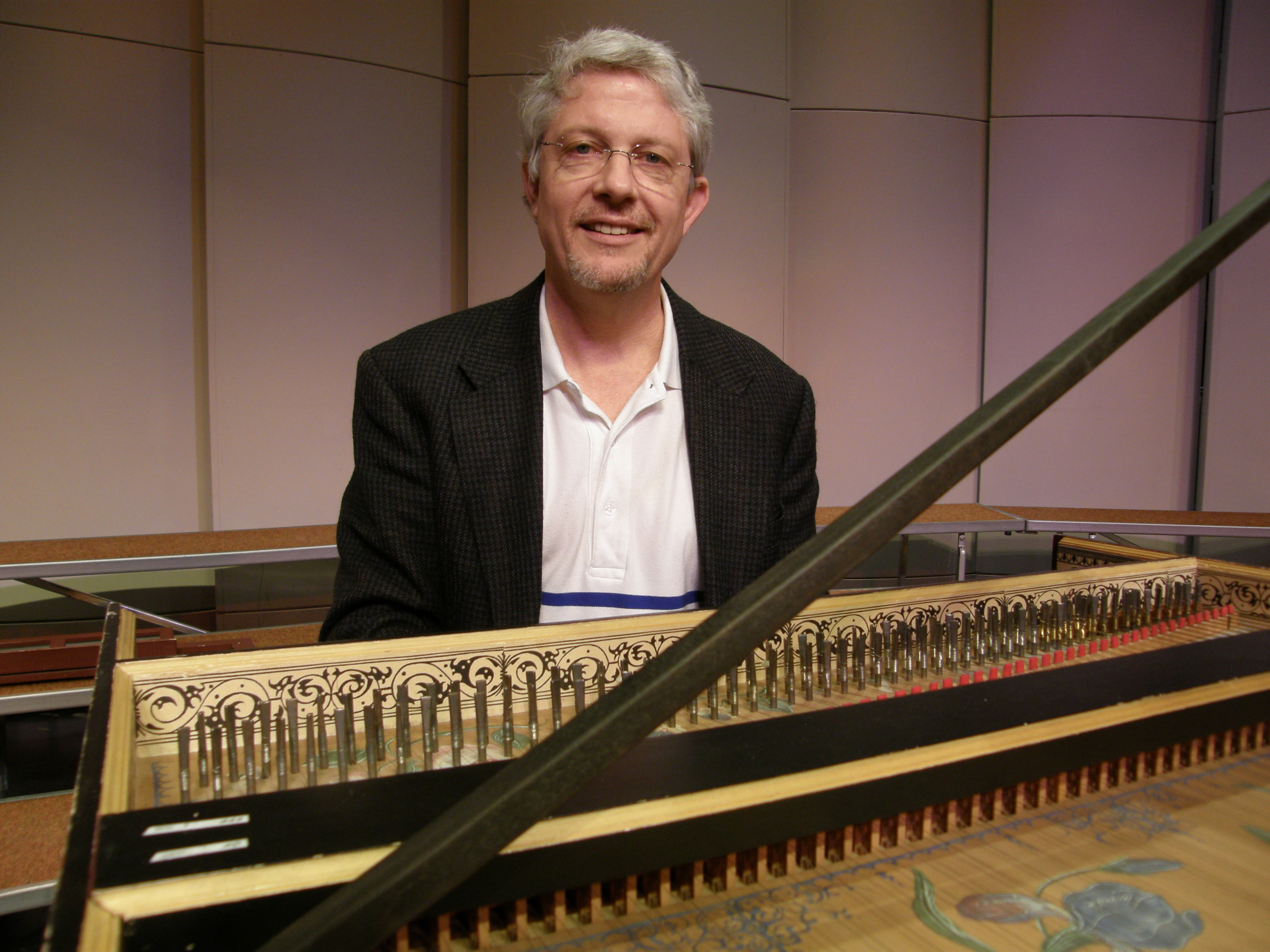 Richard Gard is seen smiling at a photograph, sitting in front of a harpsichord. 
