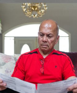 A photo shows Danilo Manimtim sitting on a couch at home looking at medical bills.