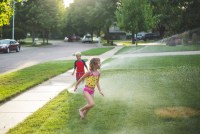 A photo shows two children in bathing suits playing in a yard with a sprinkler on a sunny day.