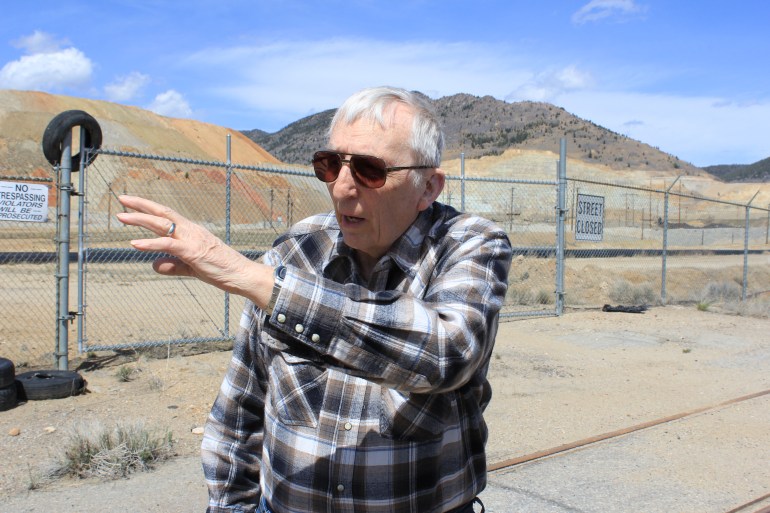 Ed Banderob points towards mounds of mine dust behind him. It is a sunny day, and the blue sky stands out against the beige landscape.