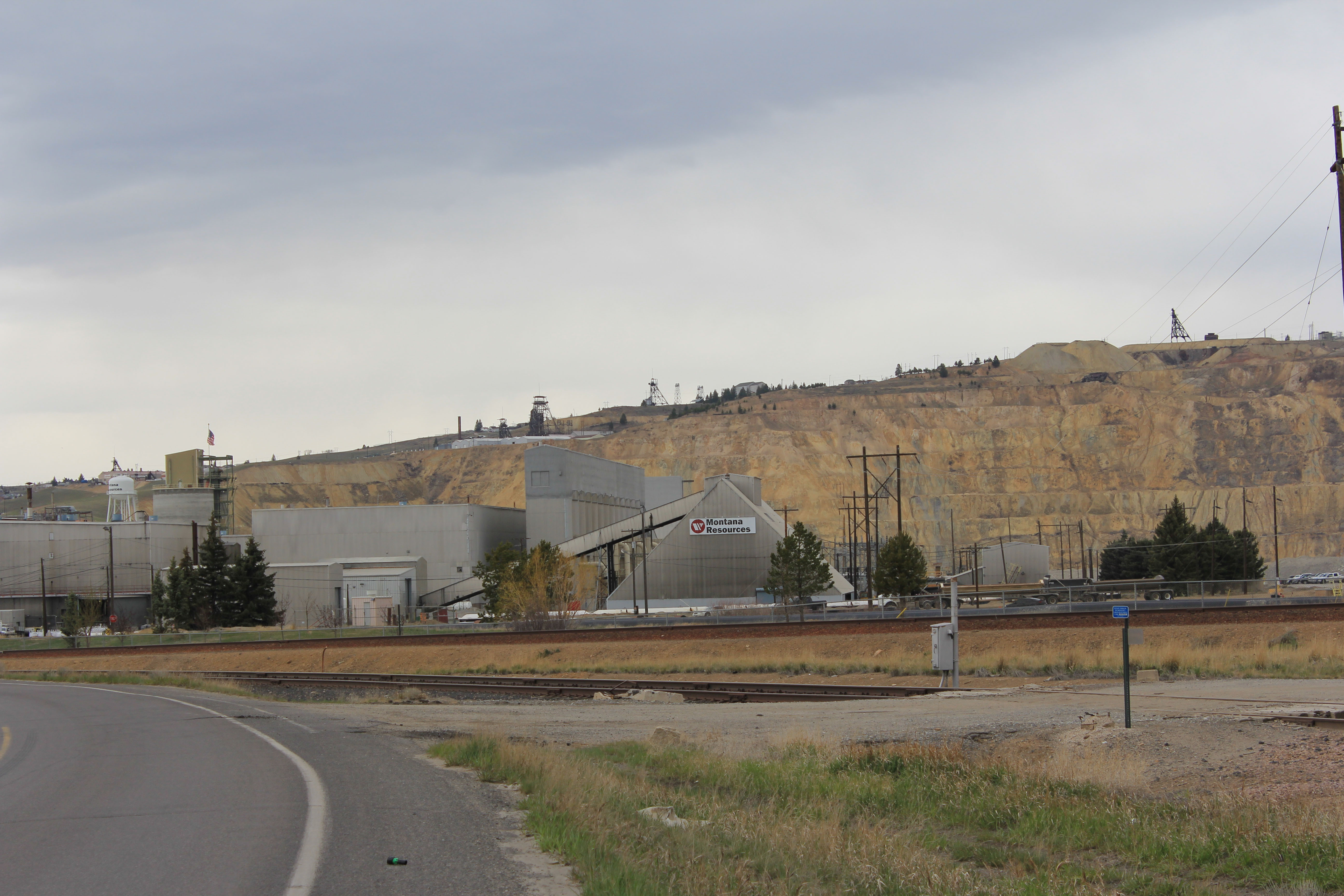 A landscape photograph of Montana Resources open-pit mine. Behind the mine are tall, sepia colored cliffs and an overcast grey sky.