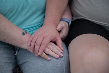 A man's hand rests on his wife's hand in her lap.