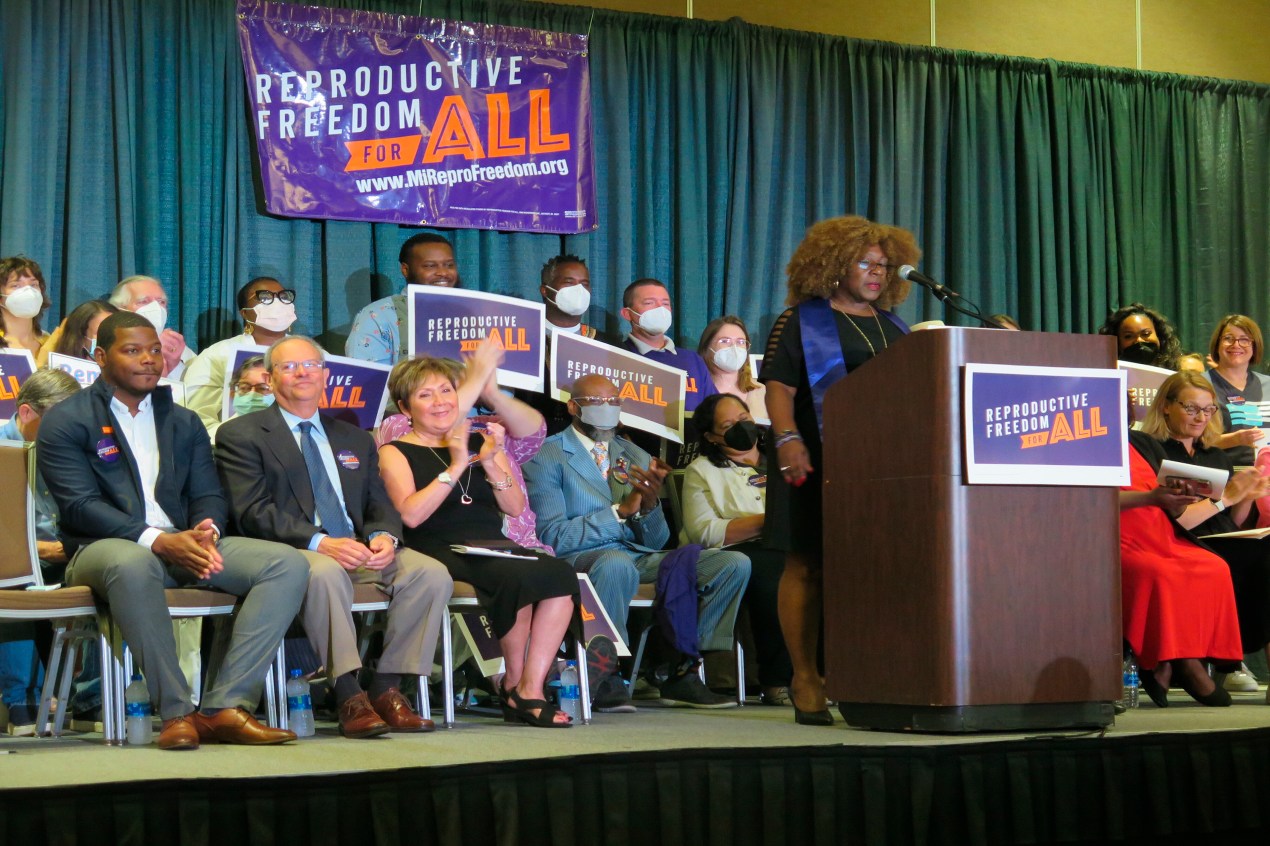 Abortion activist Leslie Mathews stands at a podium in front of a sign that reads "Reproductive Freedom For All". A group of about eight people, holding similar reading signs, are seated on the stage behind here.