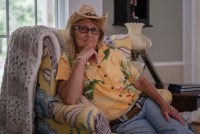 A woman in a cowboy hat and Hawaiian shirt sits in an armchair.
