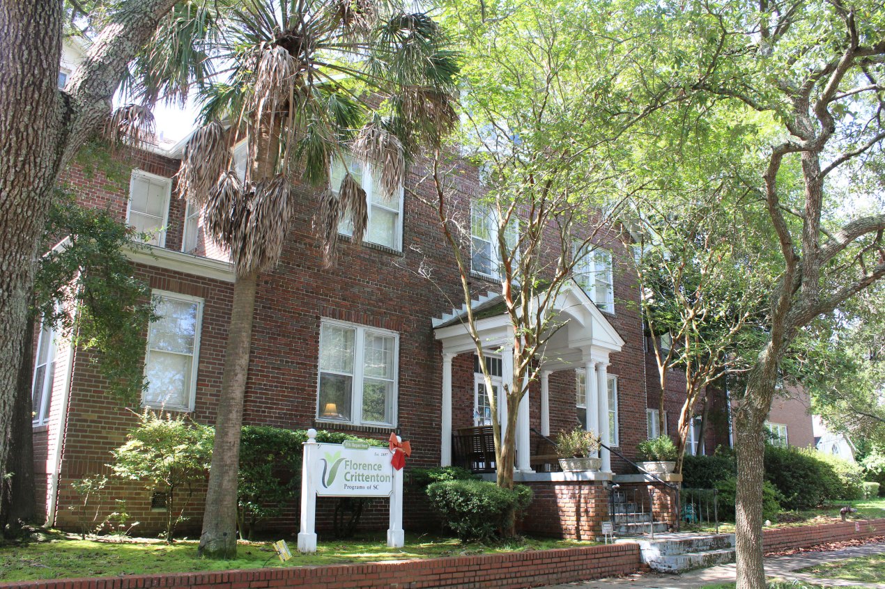 An old brick building with a white sign in front reading "Florence Crittenton Programs of SC" and surrounded by trees.