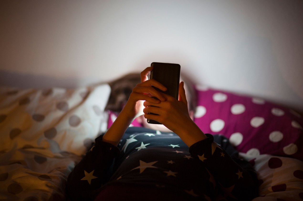 A kid lying in bed at night looking at a phone.