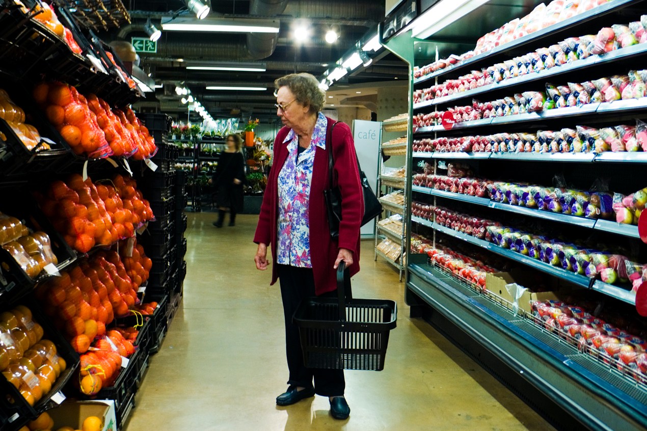 A senior woman stands in a grocery aisle holding an empty shopping basket.