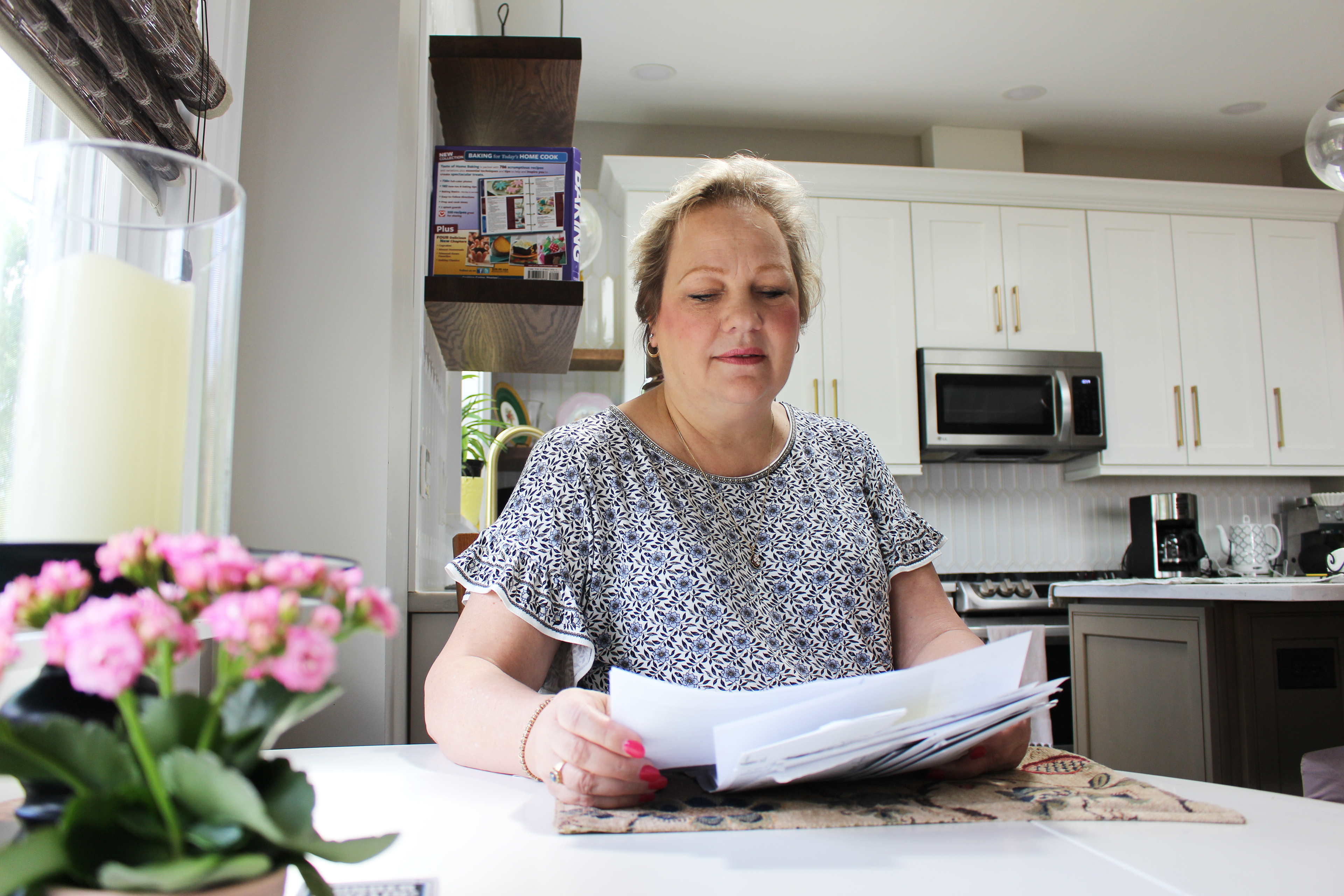 Peggy Dula is seen sitting at a kitchen table by a window. She is looking over medical bills.