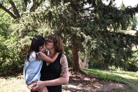 A woman holding a young child and kissing her on the cheek stands outside in front of a coniferous tree.