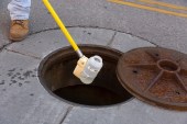 An employee collects a wastewater sample from an open manhole. A plastic bottle at the end of a long, yellow pole contains the water sample as it's lifted from the manhole.