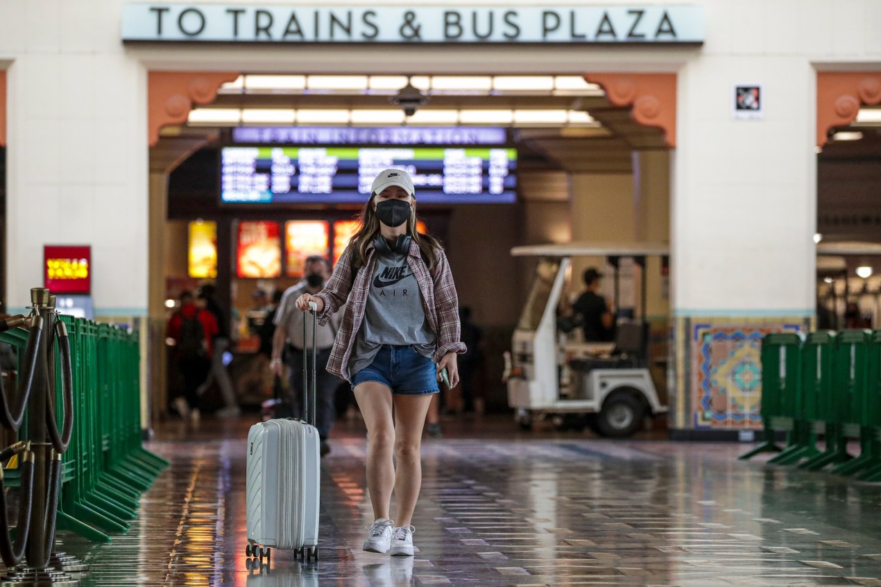 A woman a black face mask rolls a suitcase beside her. A sign behind her reads "To trains & bus plaza".