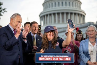 A woman in a baseball cap stands at a lecture in front of the Capitol building, surrounded by people, and holds up a phone with a man's face visible on it.
