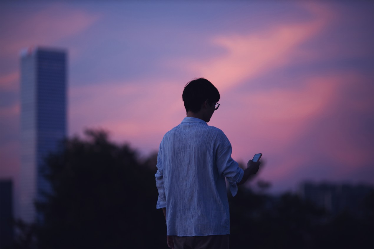 A photo shows a man from behind outside at sunset. He is looking down at his phone.