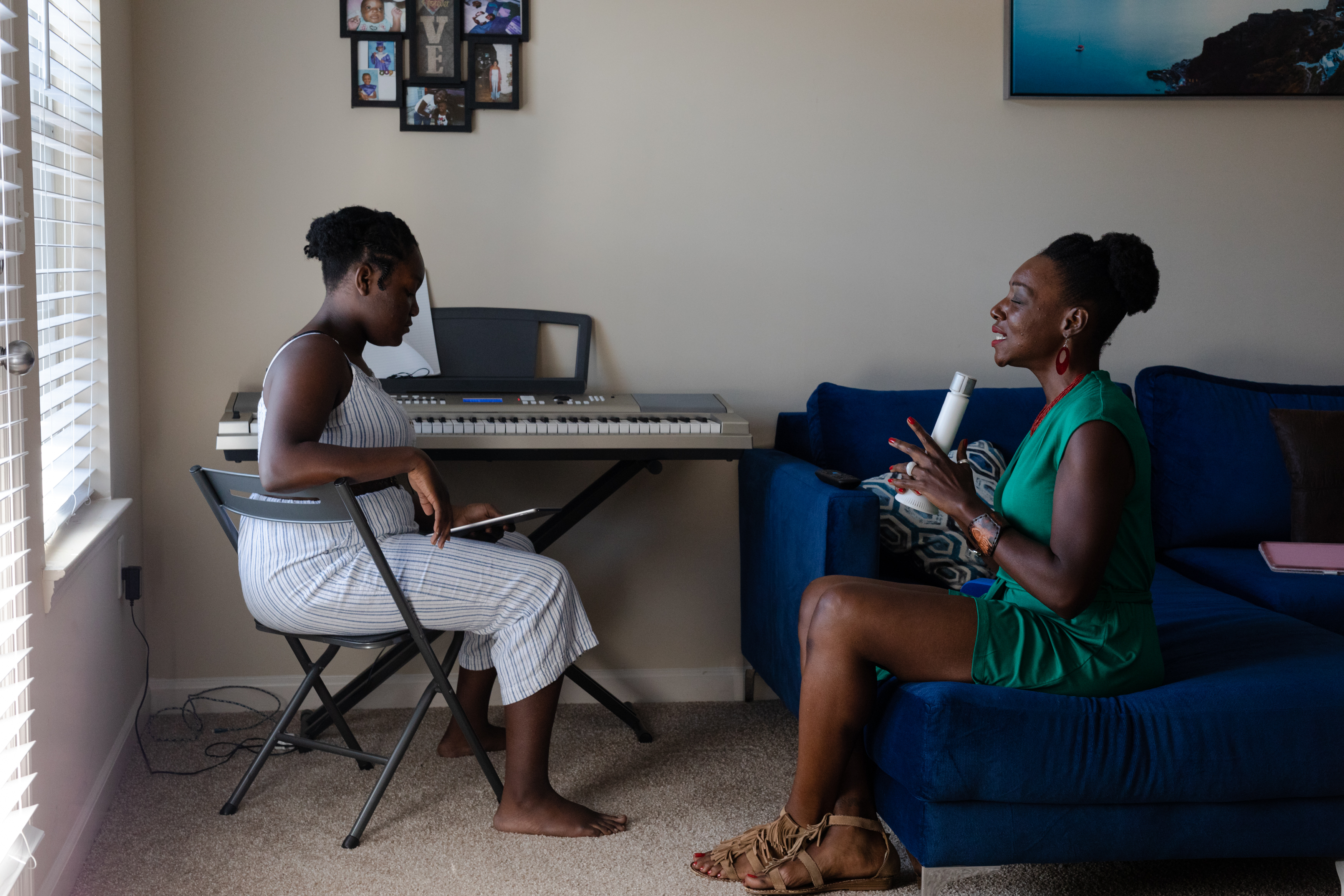 A girl and her mother sit across from each other in front of a piano. The mother sings into a microphone while the girl looks at a tablet.