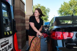A woman with wearing a black T-shirt and orange skirt is filling her car up with gas at the station. The car, to the right of the image, is black and glossy. She faces the gas pump and recoils at the price.