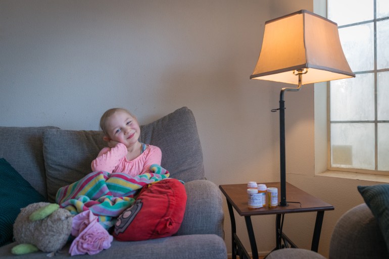 A young girl, Annabelle Lewis, wears a pink shirt and sits on a grey couch. She smiles slightly at the camera, her cheek resting against her palm. She has lost her blonde hair from chemotherapy treatments.