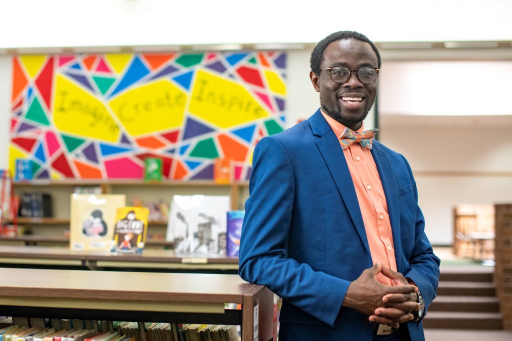 A photo shows Bertine Bahige standing inside of an elementary school library with a colorful mural on the wall behind him.