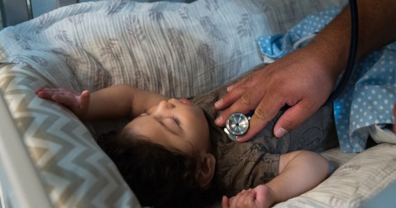 A photo shows a toddler lying asleep in bed, being checked with a stethoscope.