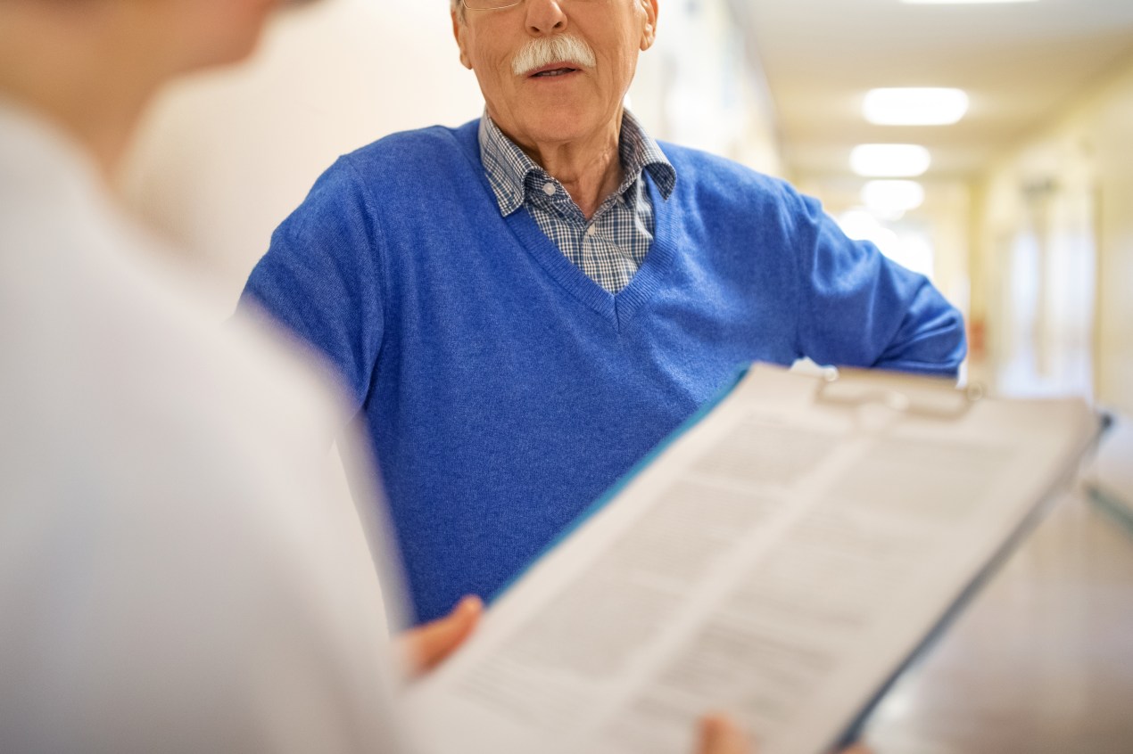 A photo shows an elderly man inside of a nursing home, talking to a medical professional, holding a clipboard with paperwork.