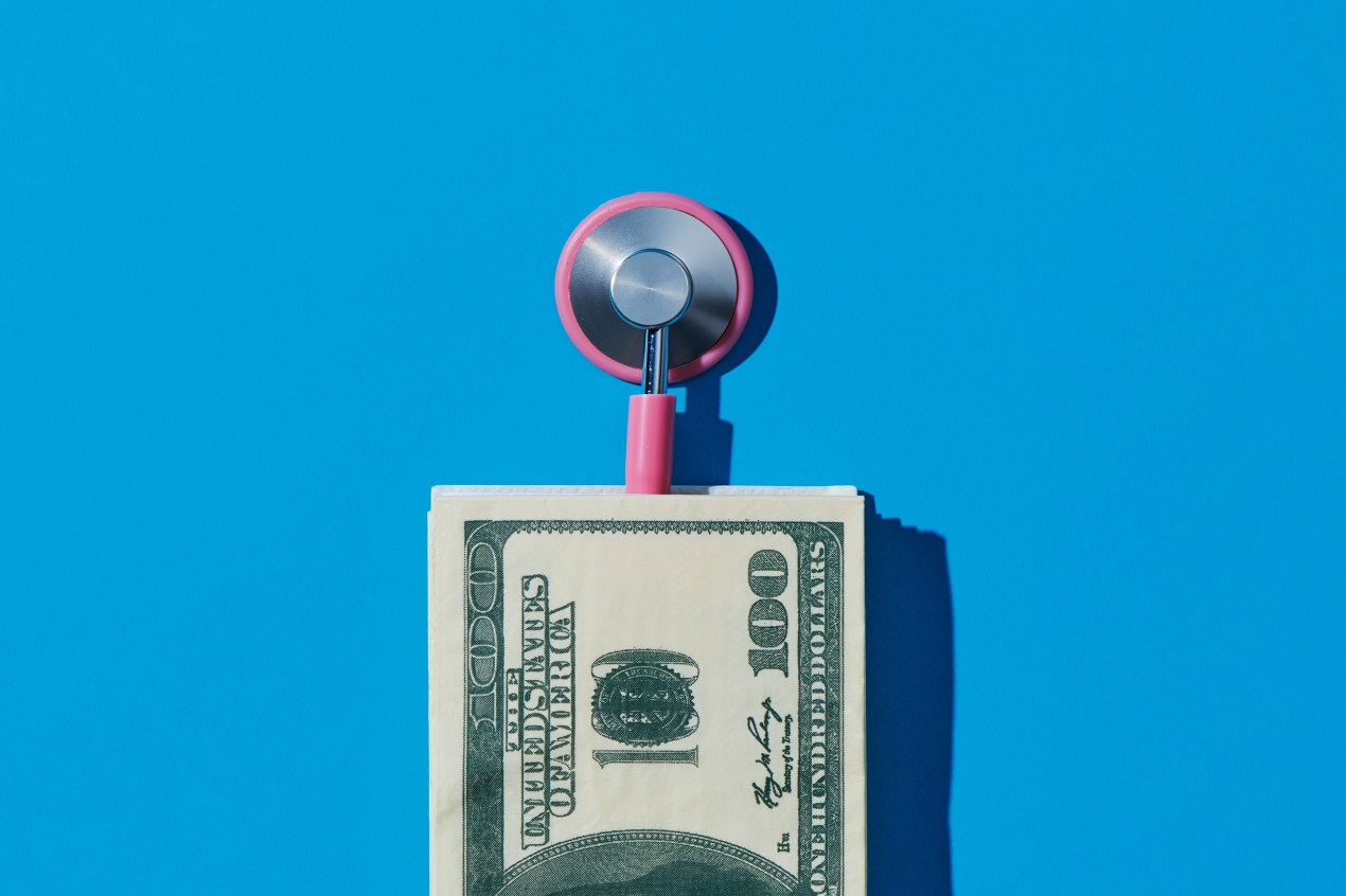 A pink stethoscope is underneath a stack of 100-dollar bills. The background is a solid blue.