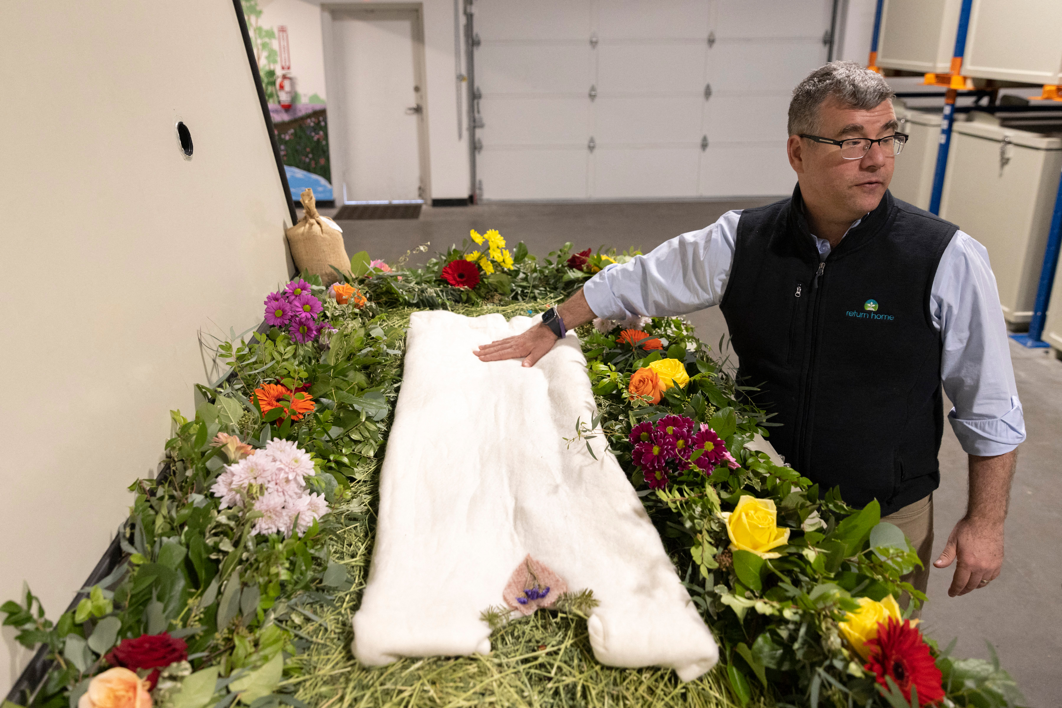 A photo shows Micah Truman standing next to a vessel decorated with flowers and compostable mementos on top of a bed of straw.