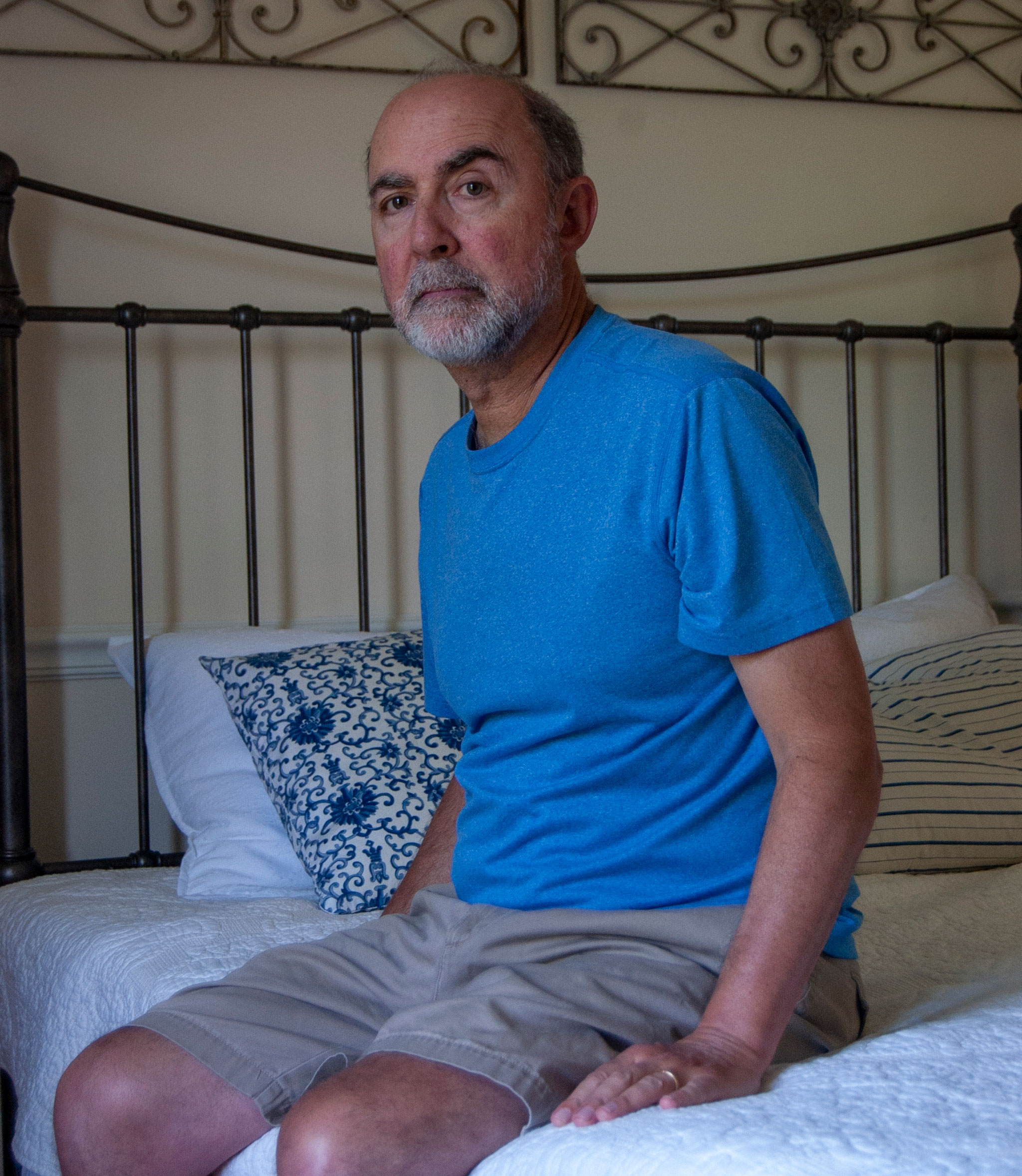 A man in a blue t-shirt sits on a bed and looks at the camera.
