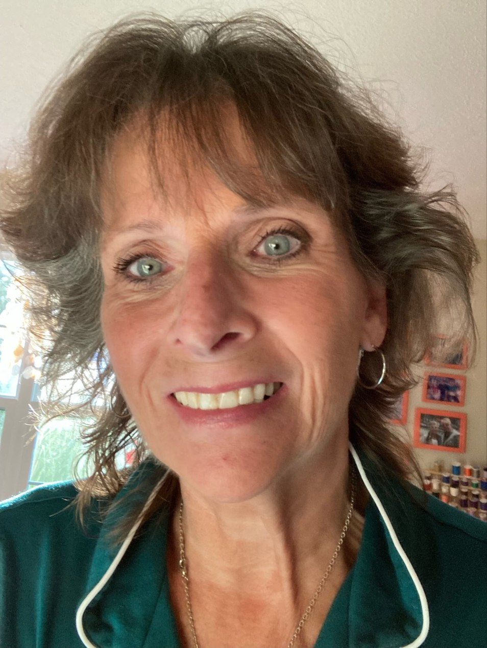 A selfie of Kelly Macauley, a woman in her 60s. She has blue eyes and brown hair with bangs, and smiles at the camera.