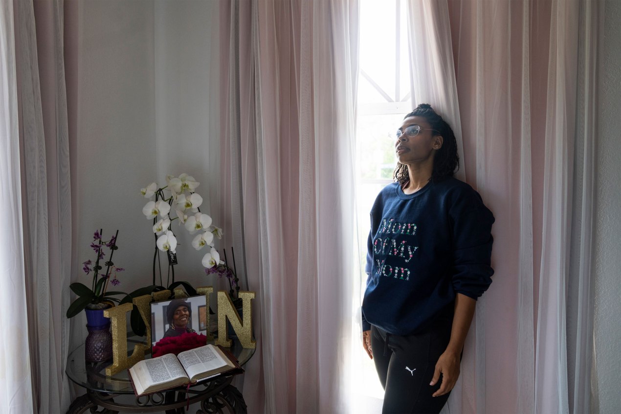 A woman stands by a window wearing a dark blue sweatshirt with the words "Mom of My Mom" on it.