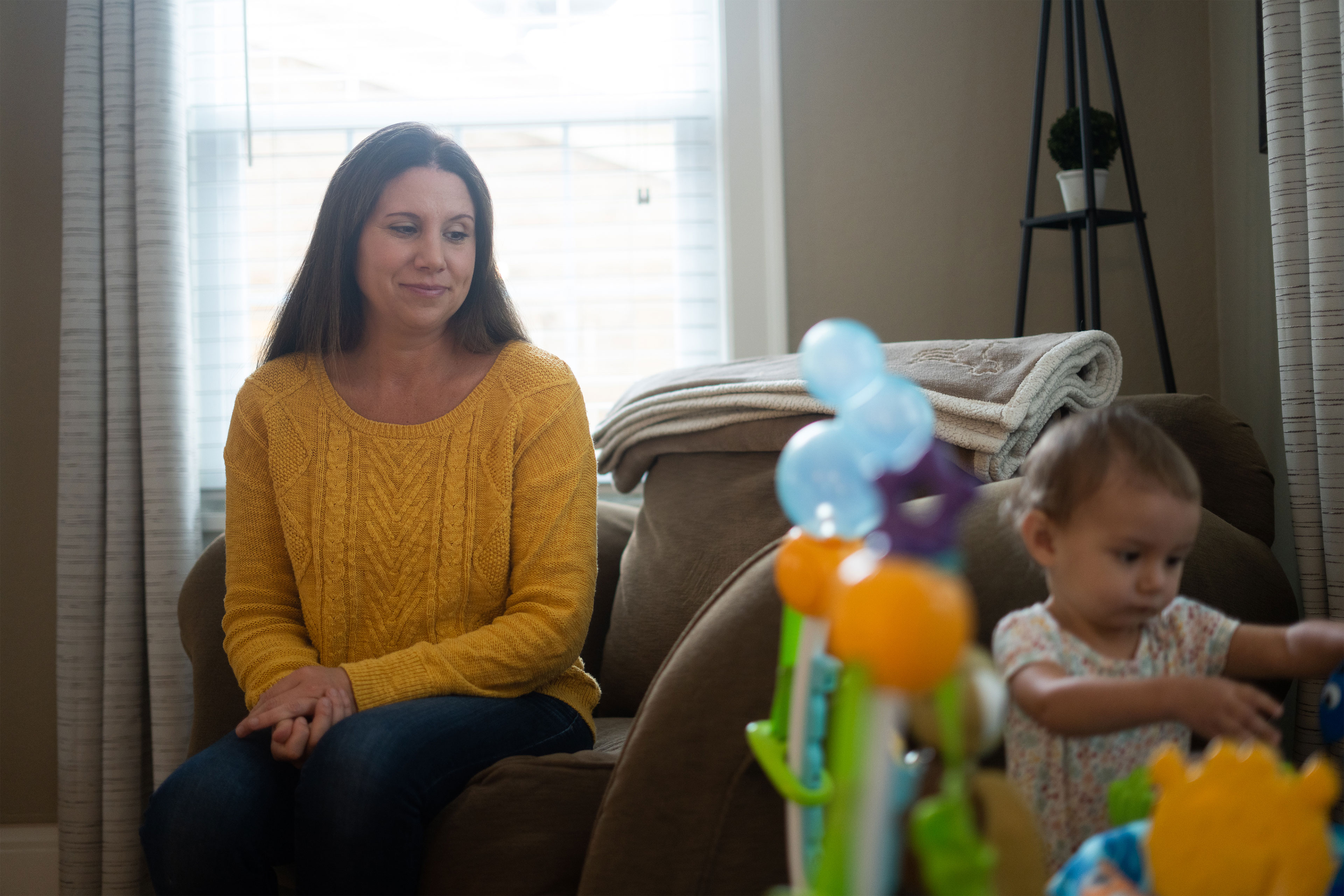 A mother with dark brown hair and a yellow sweater sits on a couch and watches her young daughter, about one year old, play with a toy.